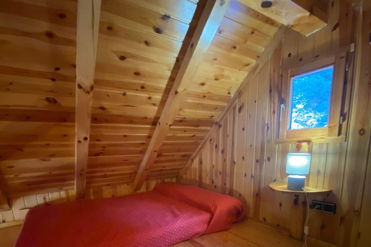 Una habitación tipo cabaña de madera con techo inclinado, un colchón rojo y una mesita de noche con lámpara.