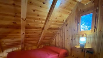 Una habitación tipo cabaña de madera con techo inclinado, un colchón rojo y una mesita de noche con lámpara.