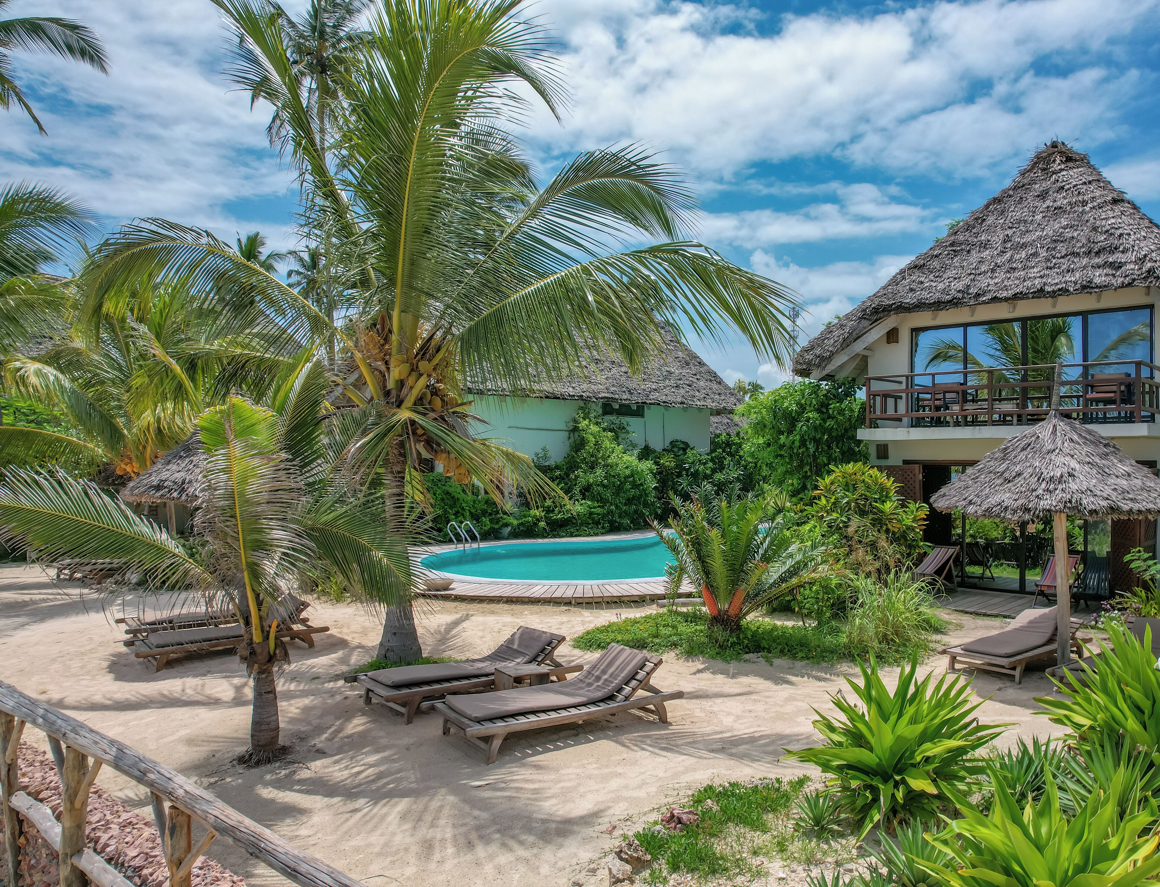 A resort with a pool, lounge chairs, palm trees, and thatched-roof buildings.