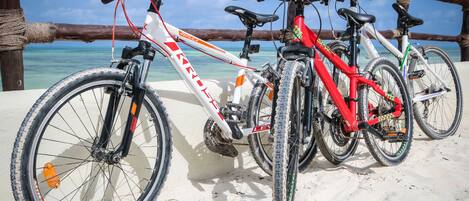 Three bicycles parked on a sandy beach with a wooden fence and ocean in the background.