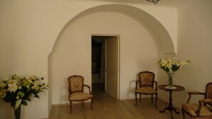 Una habitación con un arco que da a otra estancia, dos sillones tapizados, una pequeña mesa redonda con un florero, y un jarrón grande con flores a la izquierda.