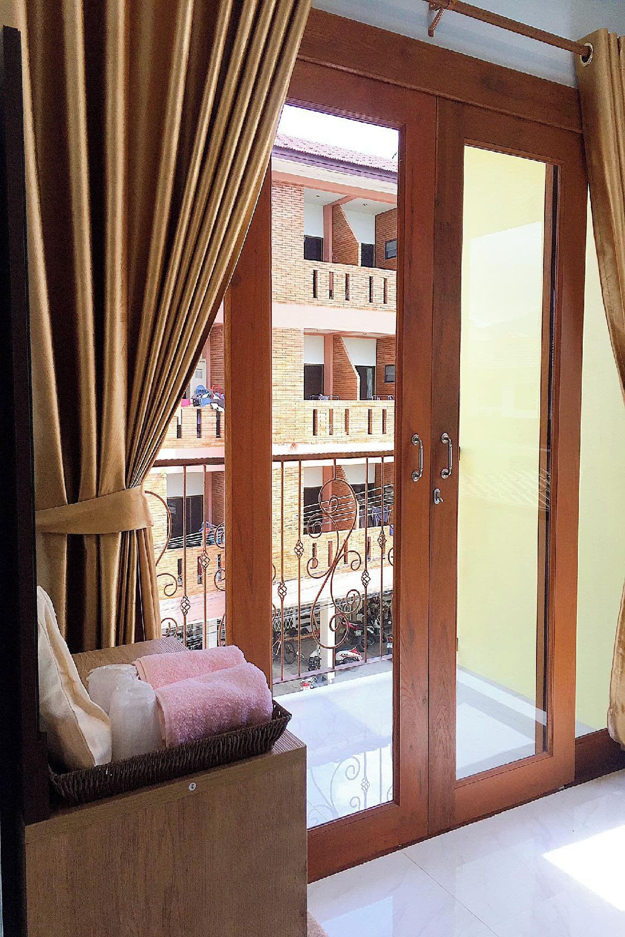 A room with a wooden door leading to a balcony, a wicker basket with towels, and a view of a building with balconies.