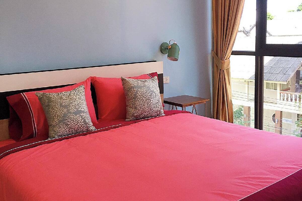 Un lit avec une couvre-lit rouge et plusieurs oreillers, une petite table et une fenêtre avec des rideaux.