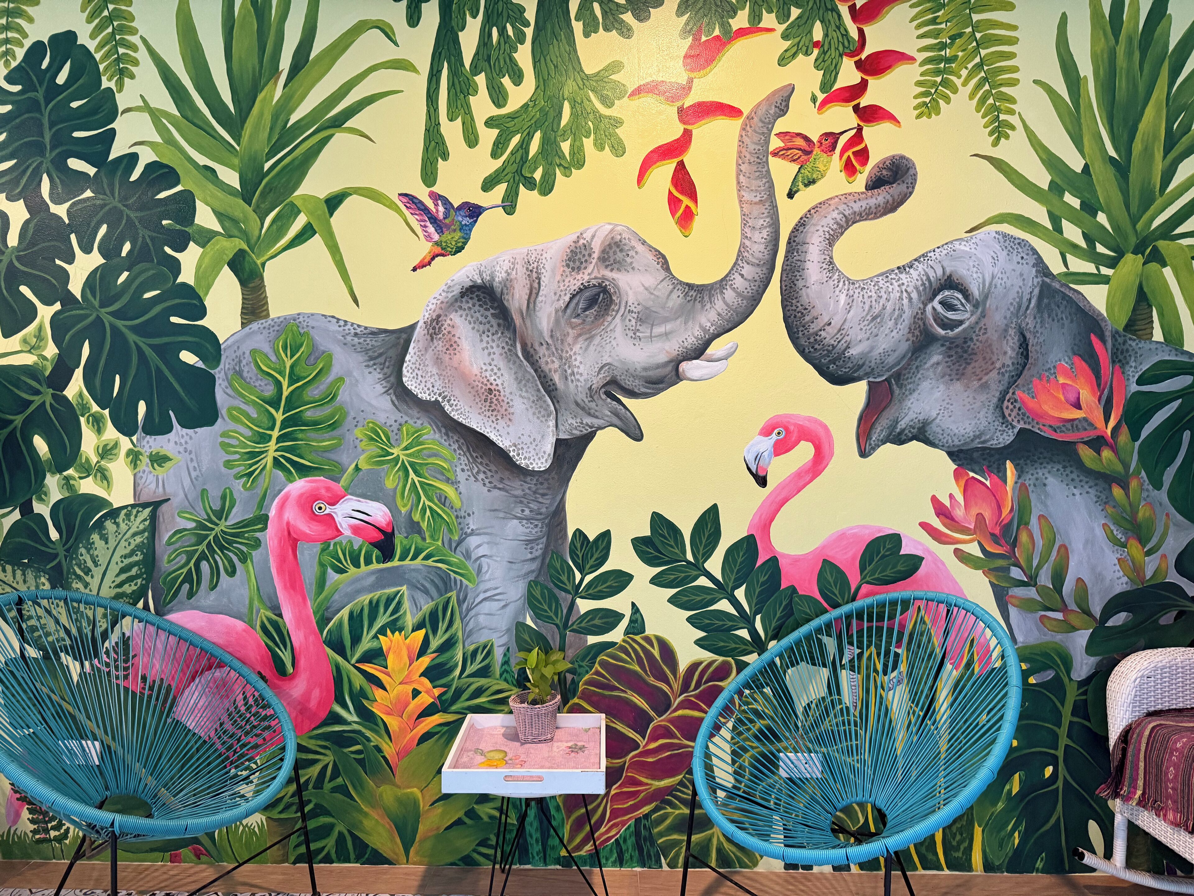 A mural with elephants, flamingos, and tropical foliage. There are two blue chairs in front of the mural.