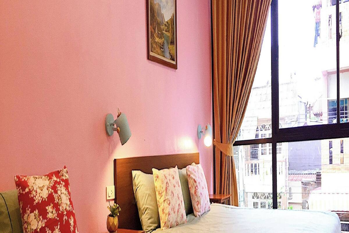 Une chambre d’hôtel avec deux lits, des murs roses, un tableau, une table de chevet et une fenêtre avec des rideaux.