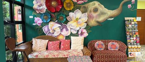 A room with a large floral mural, a wicker sofa, patterned cushions, and a tiled floor.