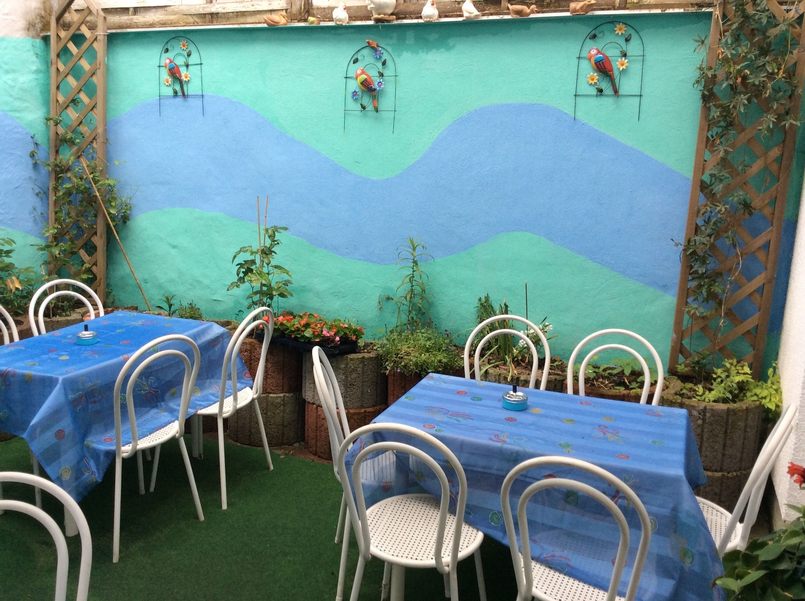Une terrasse extérieure avec des tables et des chaises, un mur peint orné de motifs ondulés et des figurines d’oiseaux décoratives.