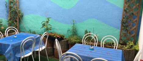 Une terrasse extérieure avec des tables et des chaises, un mur peint orné de motifs ondulés et des figurines d’oiseaux décoratives.