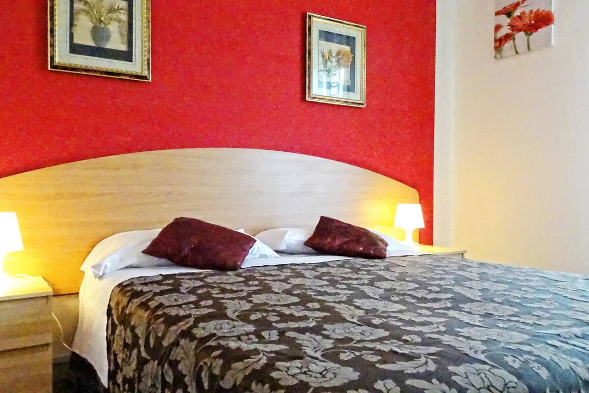 Un lit bien fait, recouvert d’une couvre-lit à motifs floraux et agrémenté de deux oreillers, de part et d’autre duquel se trouvent des tables de chevet avec des lampes. La pièce est décorée de murs rouges et de tableaux encadrés qui ornent le mur au-dessus du lit.