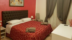 Habitación de hotel con pared roja, cama grande con cabecera capitonada, un cuadro sobre la cama, una silla, un escritorio y una cortina.
