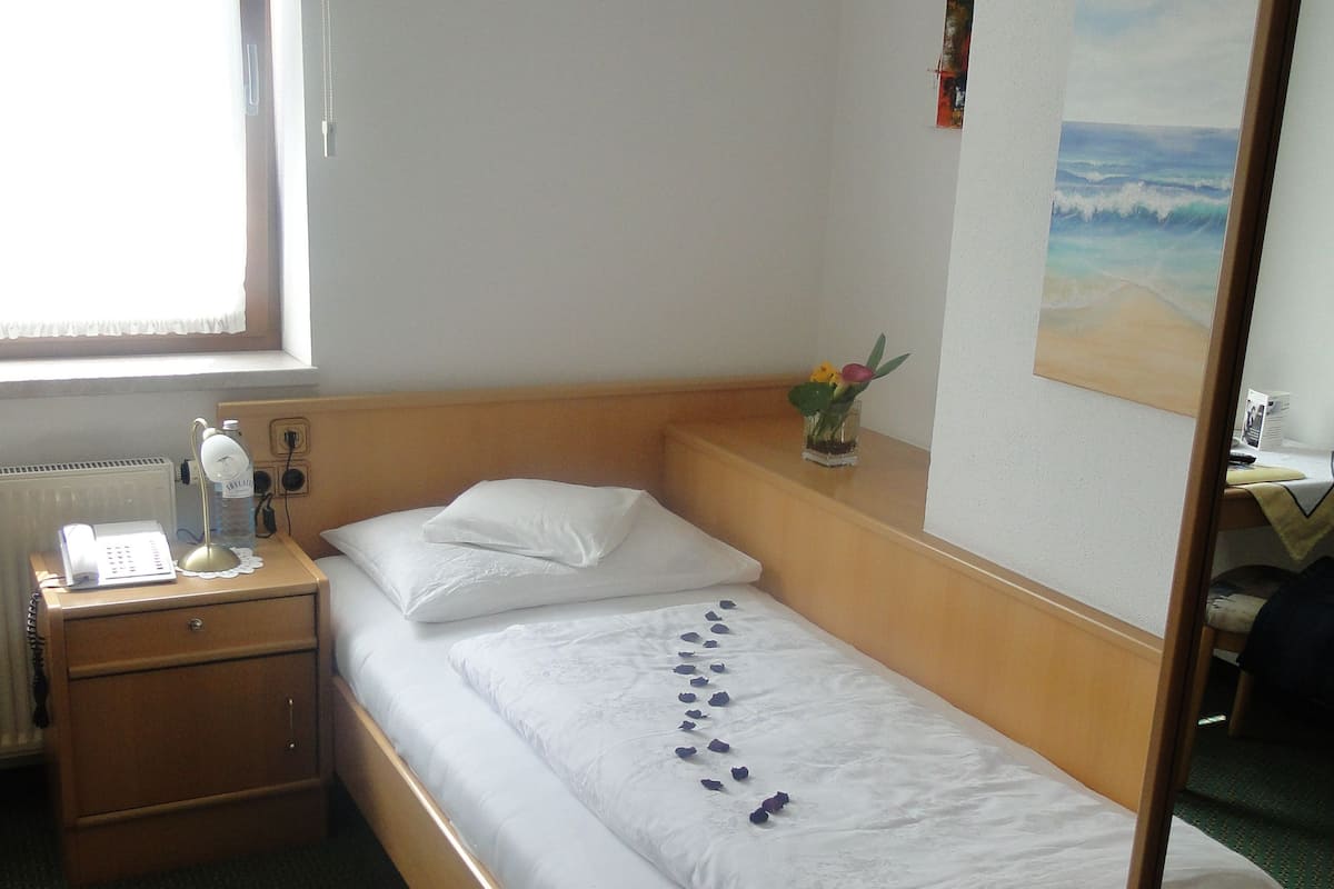 A single bed with white linens, a bedside table with a lamp, a small vase with flowers, and a painting on the wall.