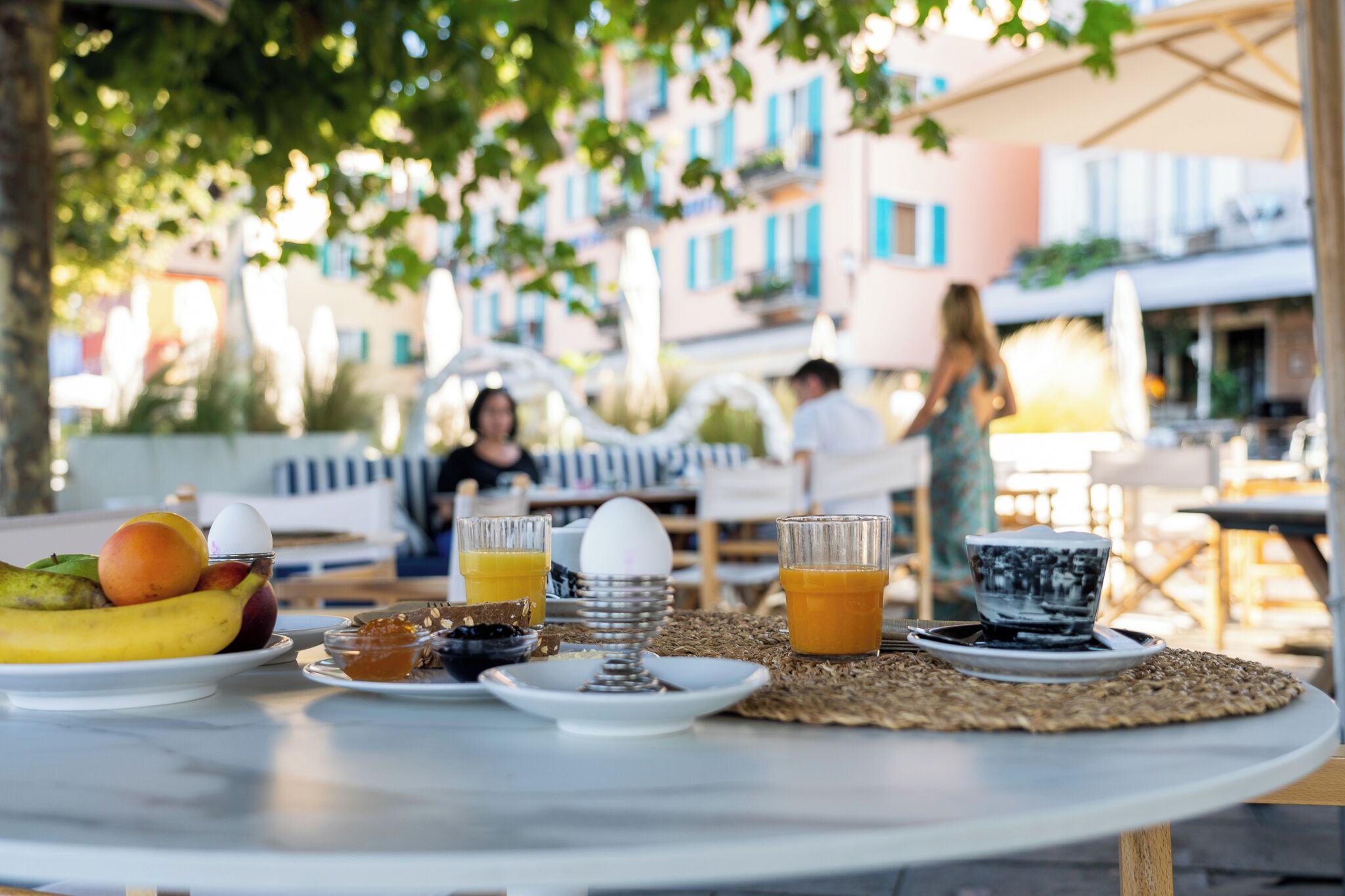 A breakfast table with fruits, eggs, and beverages, set outdoors with buildings in the background.