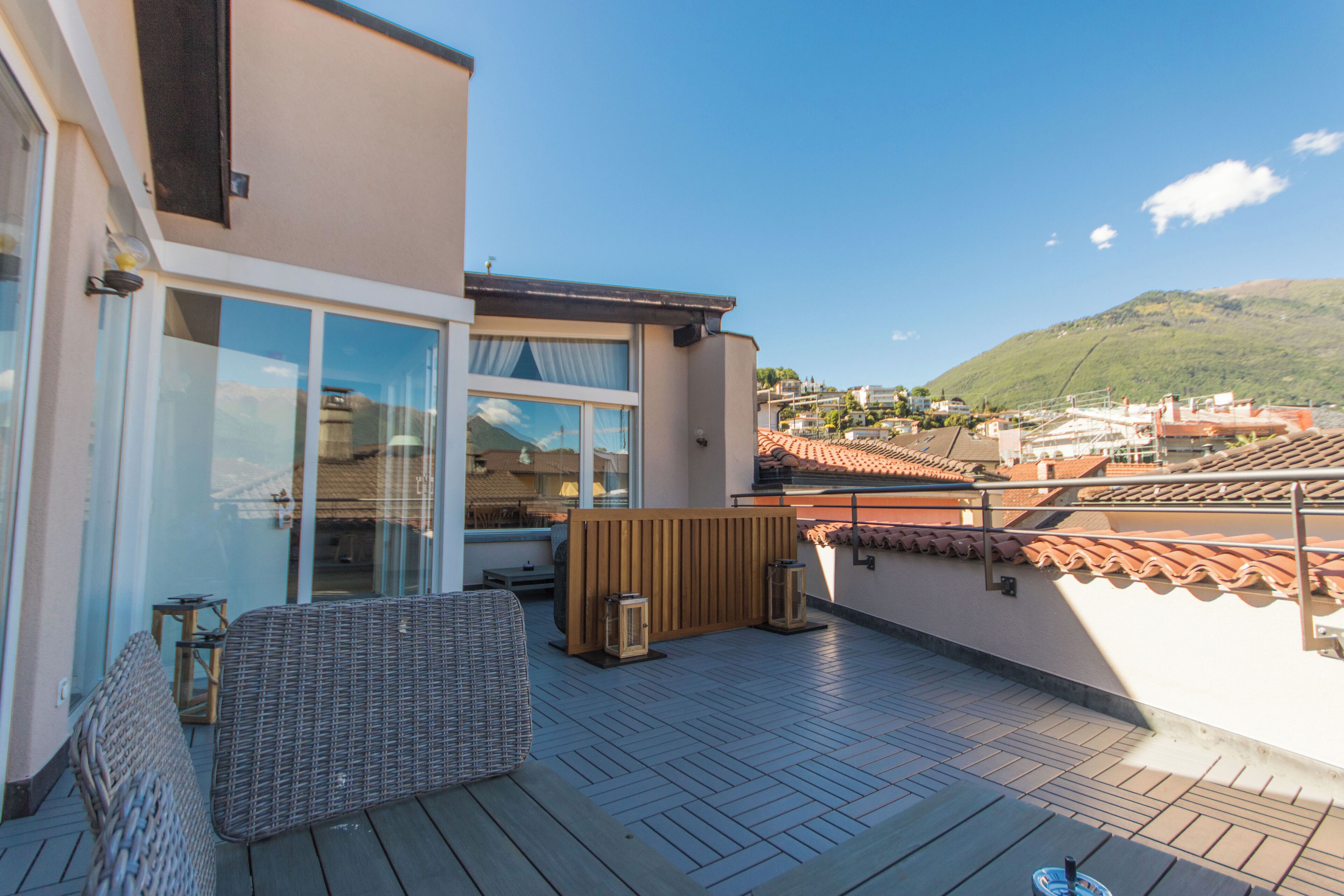 A rooftop terrace with a wooden deck, a seating area with a wicker chair, and a view of a town and hills in the background.