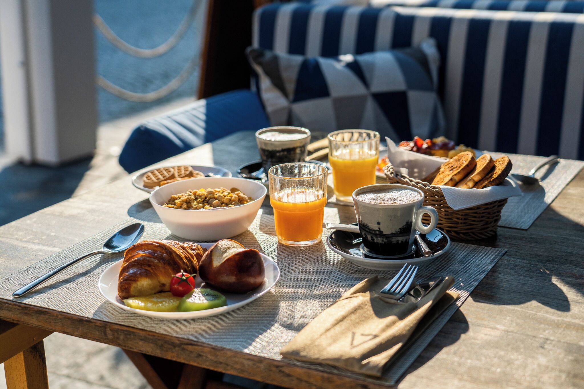 A breakfast setup on a wooden table with croissants, fruits, a bowl of cereal, glasses of orange juice, and a cup of coffee.