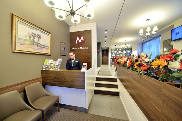 A hotel reception desk with a wooden counter, a framed painting on the wall, and a row of colorful flowers on the right.