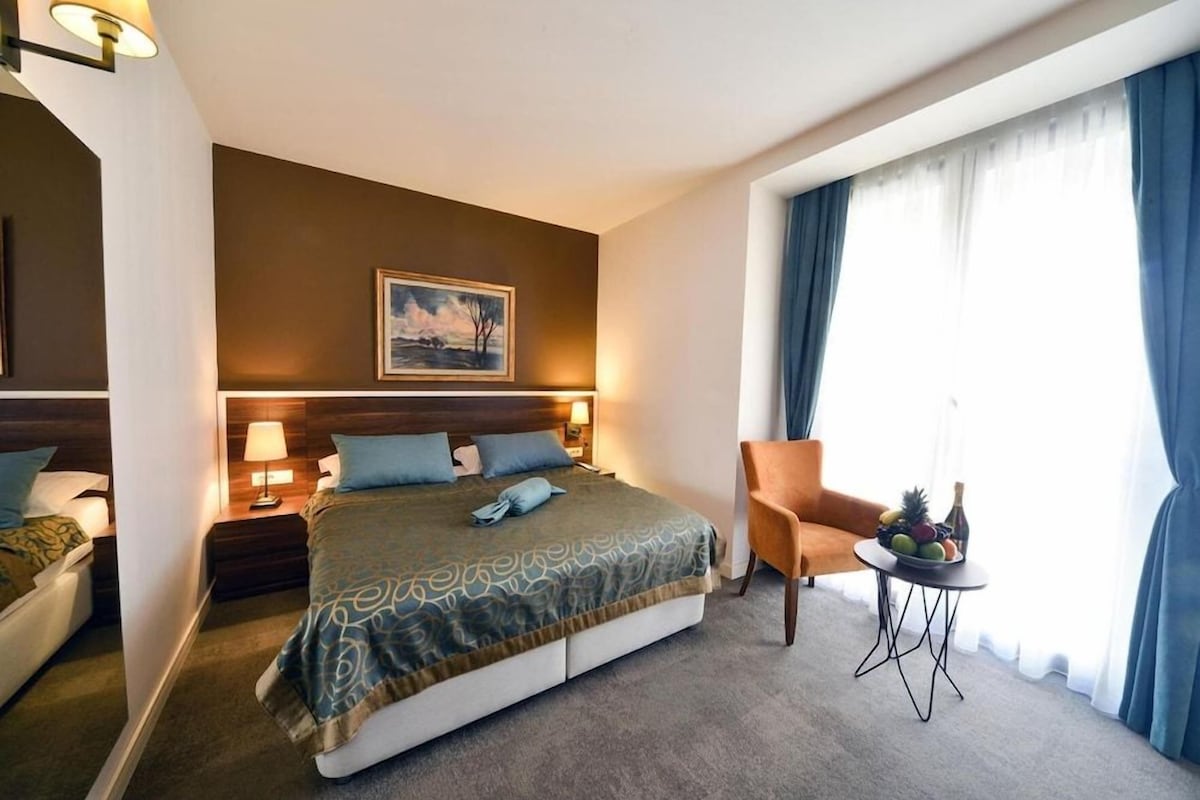 A hotel room with a large bed, two bedside tables with lamps, a chair, a small table with fruit, and a painting on the wall.