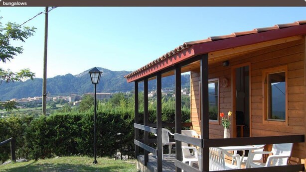 A wooden cabin with a porch, white plastic chairs, and a view of mountains. - Camping Morunys (Sant Llorenc de Morunys)