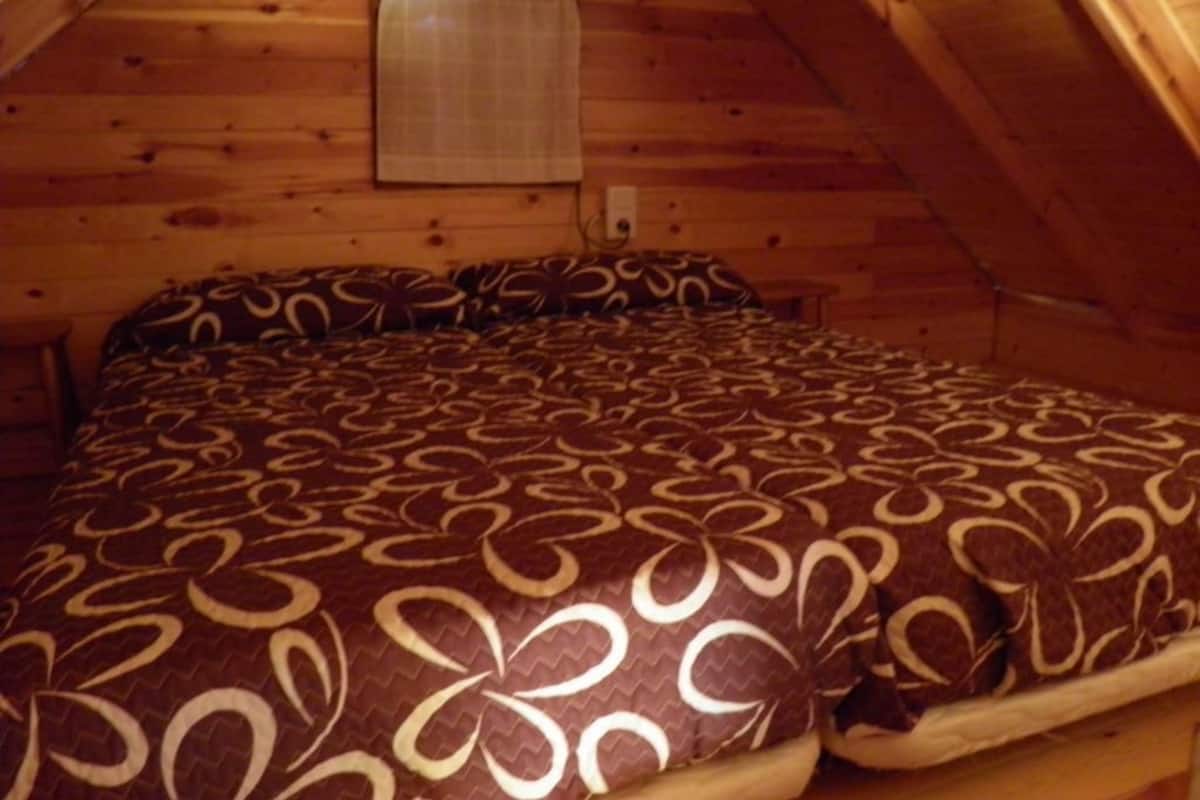 Un dormitorio acogedor en el ático con techo inclinado de madera, una cama con ropa de cama estampada y una lámpara fijada a la pared.