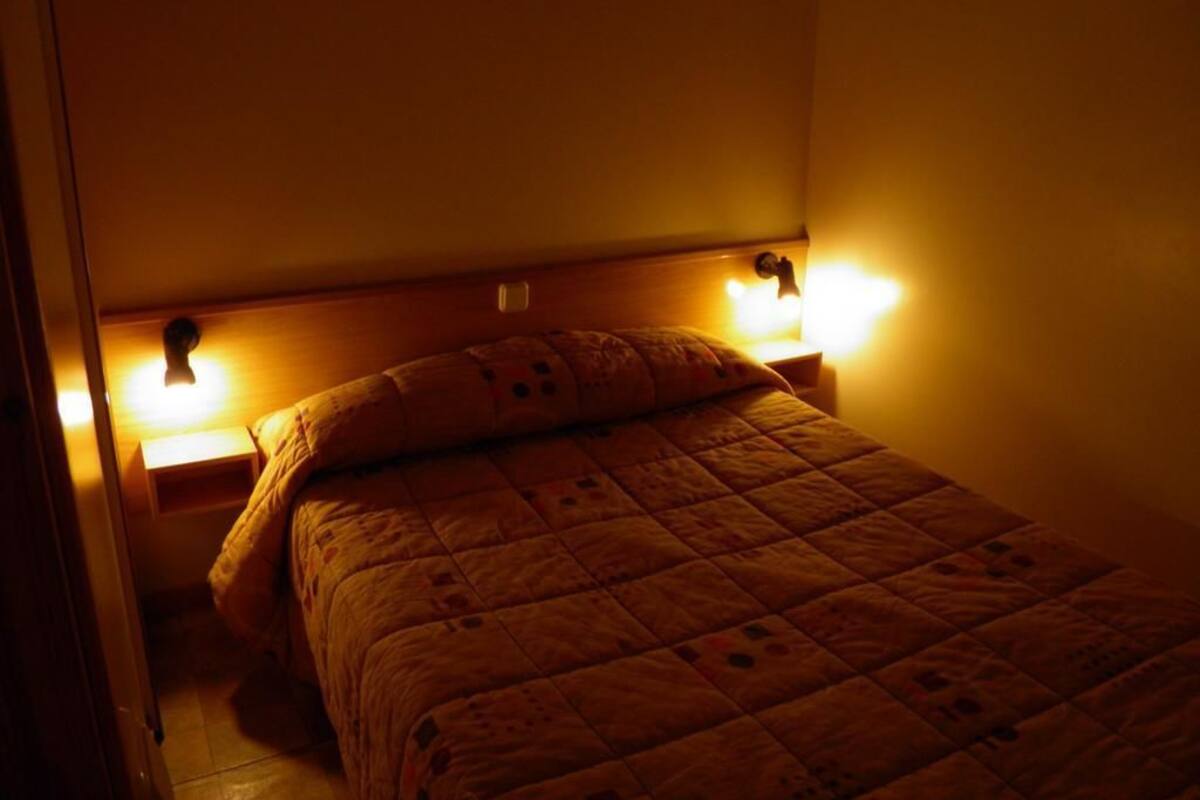 Una cama individual con colcha, dos lámparas de noche y un cabecero de madera en una habitación con poca luz.
