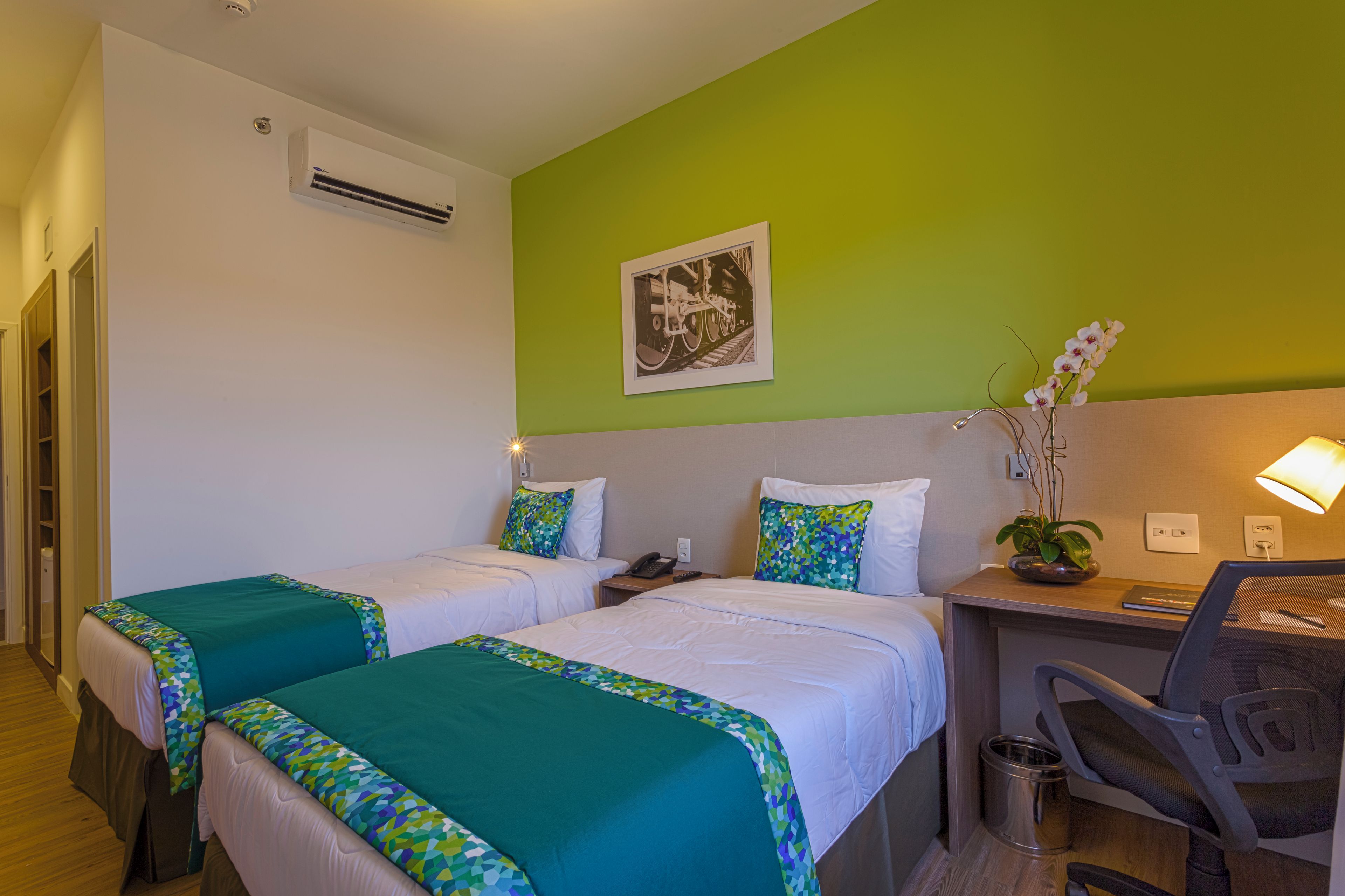 A hotel room with two beds, a desk, a chair, a trash bin, and a wall-mounted air conditioner.