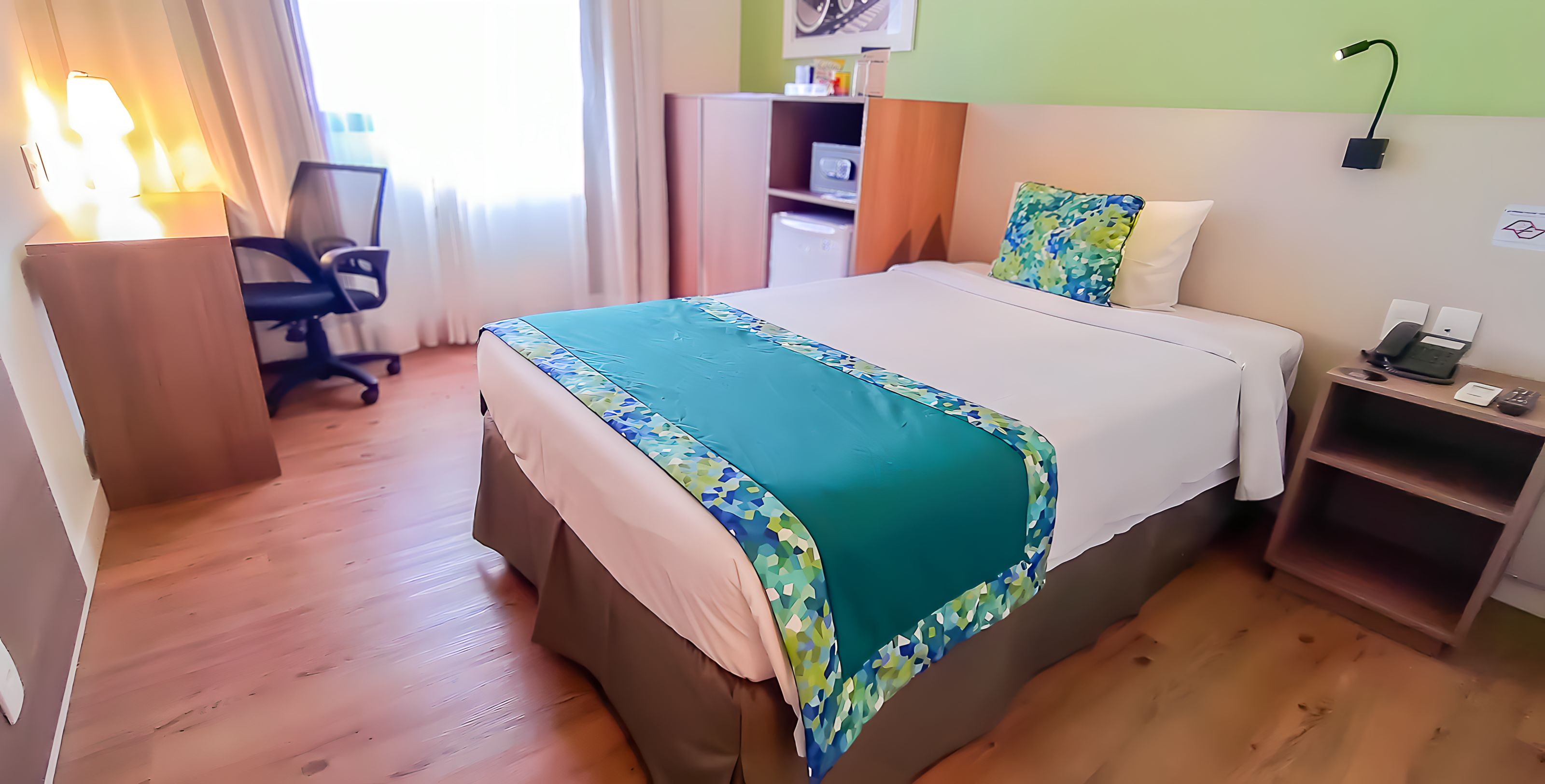 A hotel room with a bed, desk, chair, and a small shelf with a lamp.