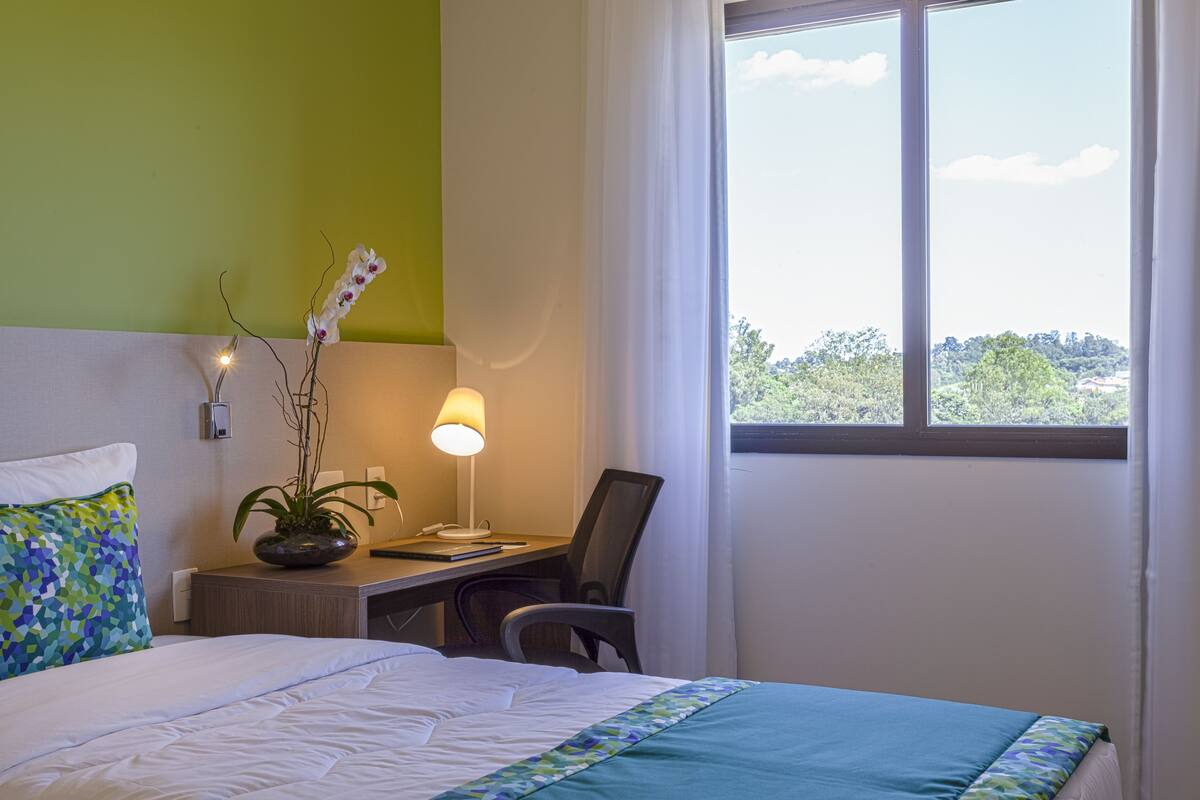 Een hotelkamer met een bed, bureau, stoel, lamp en raam met uitzicht op groen.