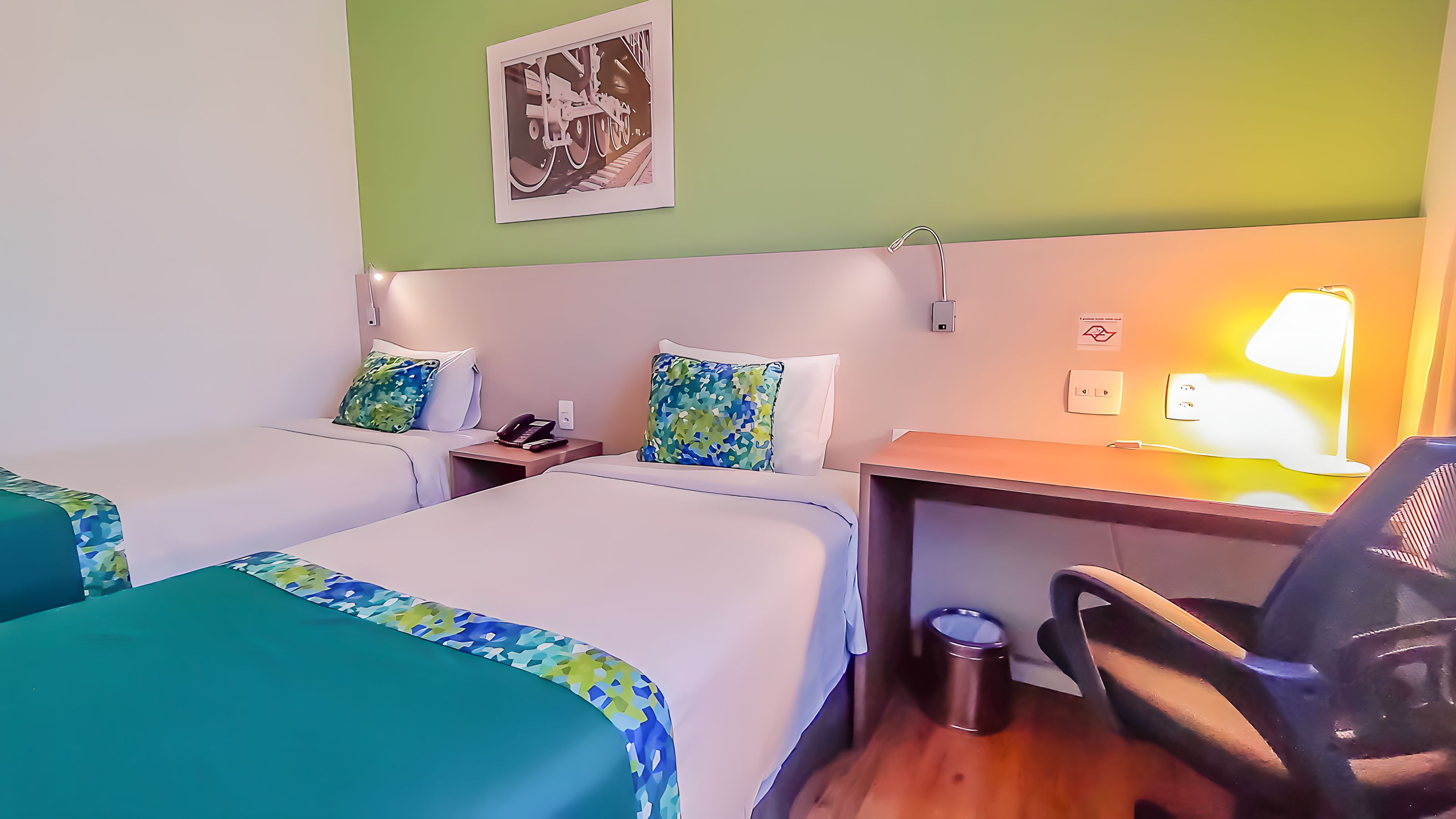 A hotel room with two beds, a desk, a chair, a lamp, and a trash bin.