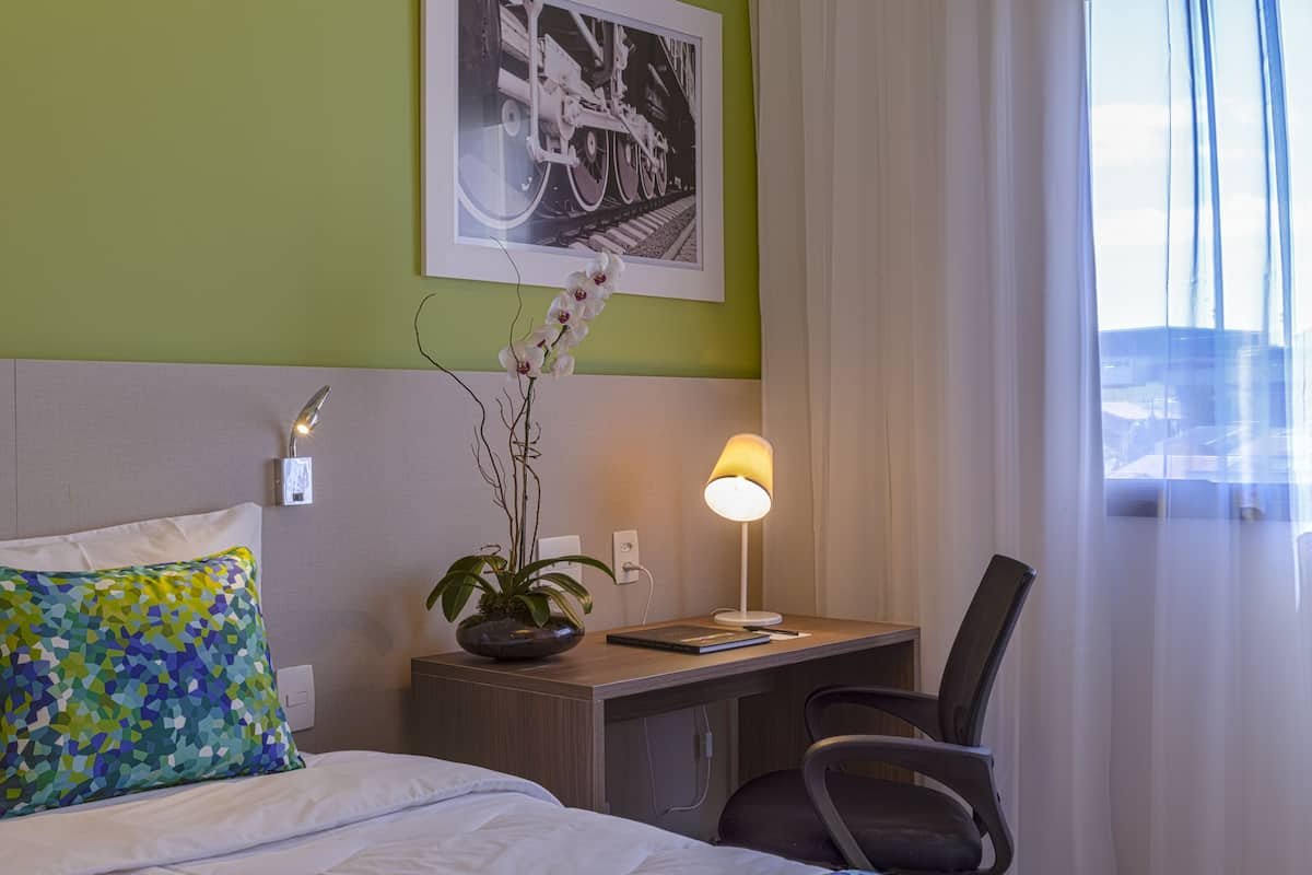 Een hotelkamer met een bed, een bureau, een stoel, een lamp, een plant en een lijst met foto op de muur.