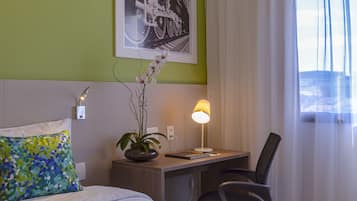 A hotel room with a bed, a desk, a chair, a lamp, a plant, and a framed picture on the wall.