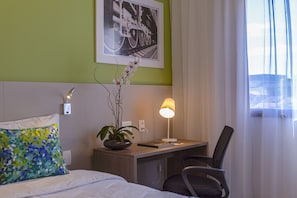 Habitación de hotel con cama, escritorio, silla, lámpara, planta y un cuadro en la pared.
