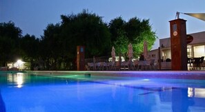 A swimming pool with lounge chairs and umbrellas, surrounded by trees and a building with a clock tower.