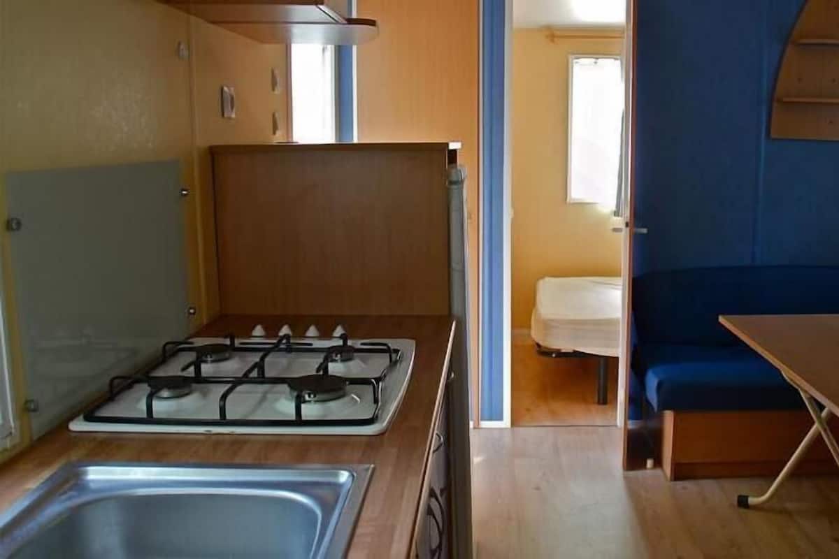Pieni, käytännöllinen keittiönurkkaus, jossa on kaasuliesi, pesuallas sekä pieni ruokailutila, jossa on pöytä ja penkki.