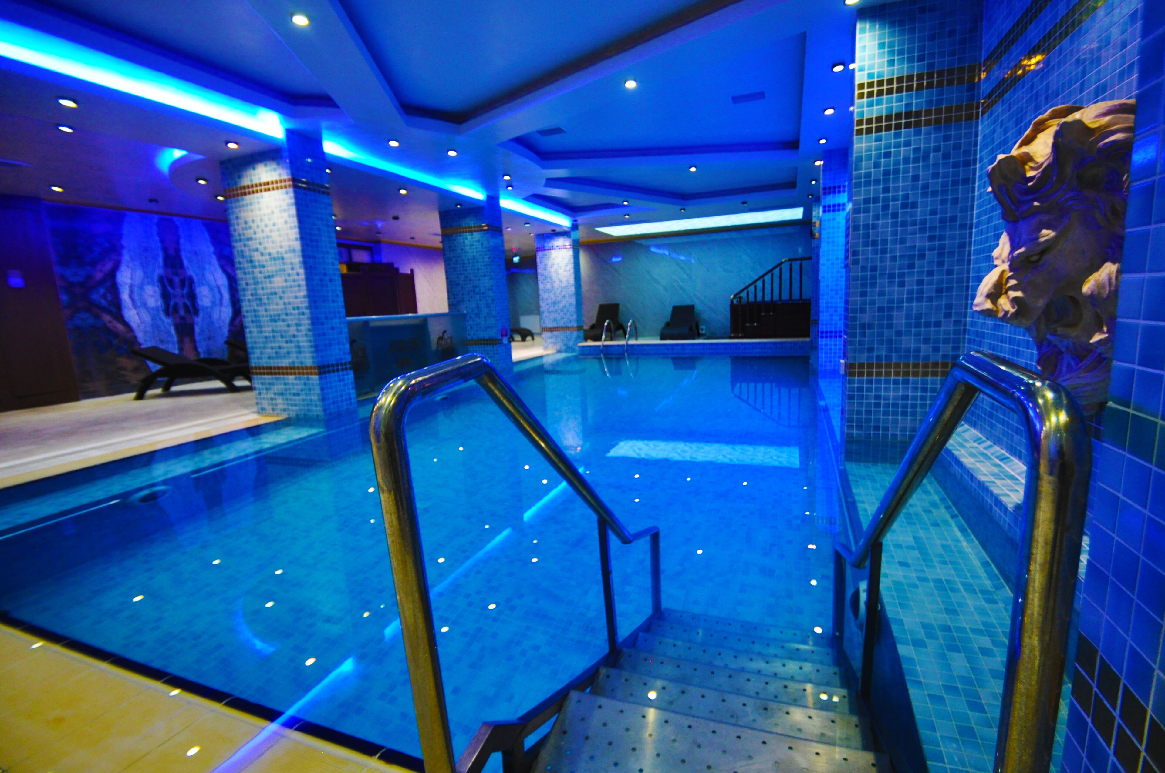 A swimming pool with blue lighting, a staircase, and a mosaic wall.
