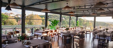 Un comedor interior con mesas y sillas, ventanas amplias y lámparas colgantes.