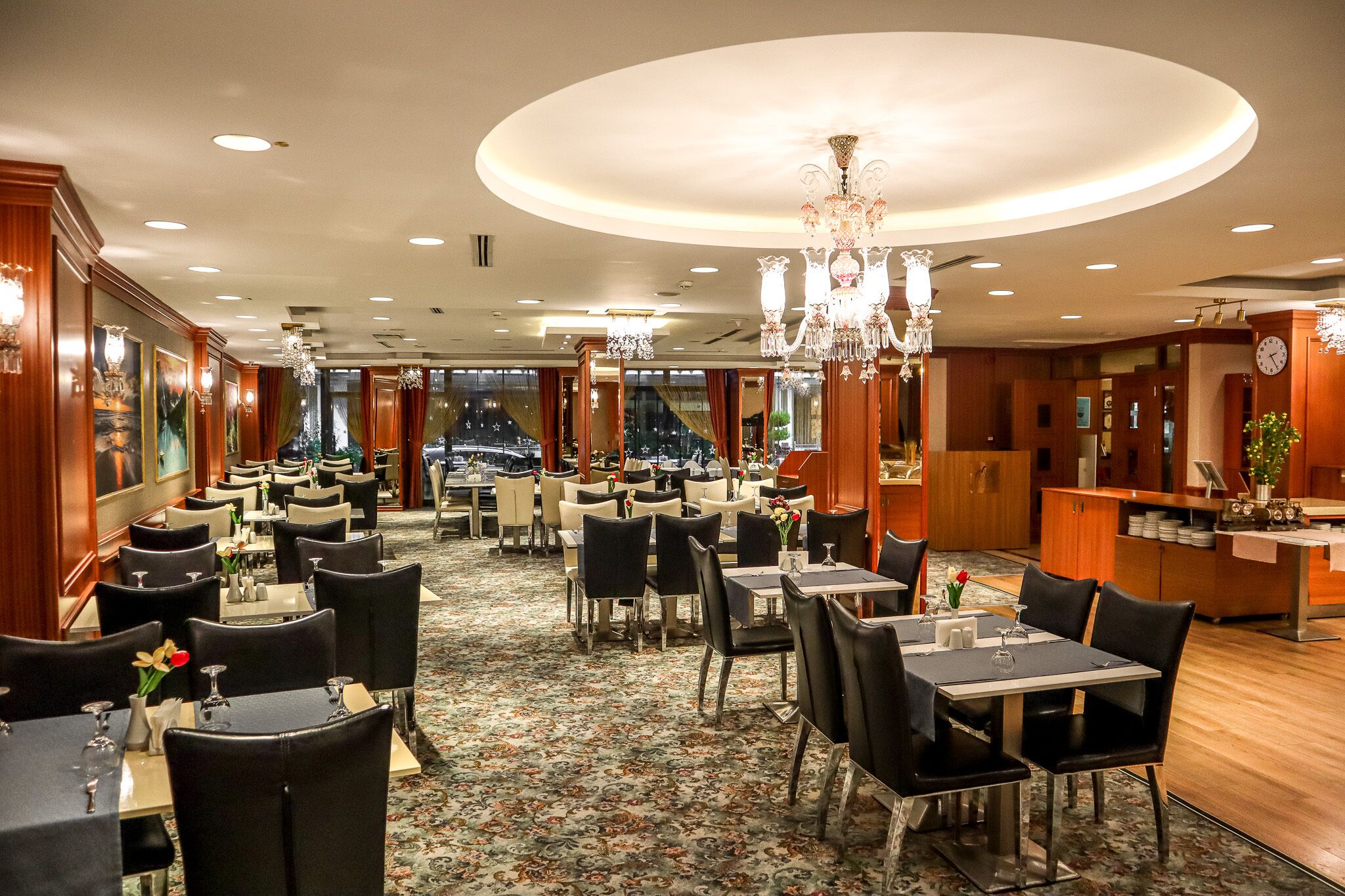 Ein geräumiges Restaurant mit mehreren Tischen und Stühlen, eleganten Kronleuchtern und Blick auf die Skyline durch große Fenster.