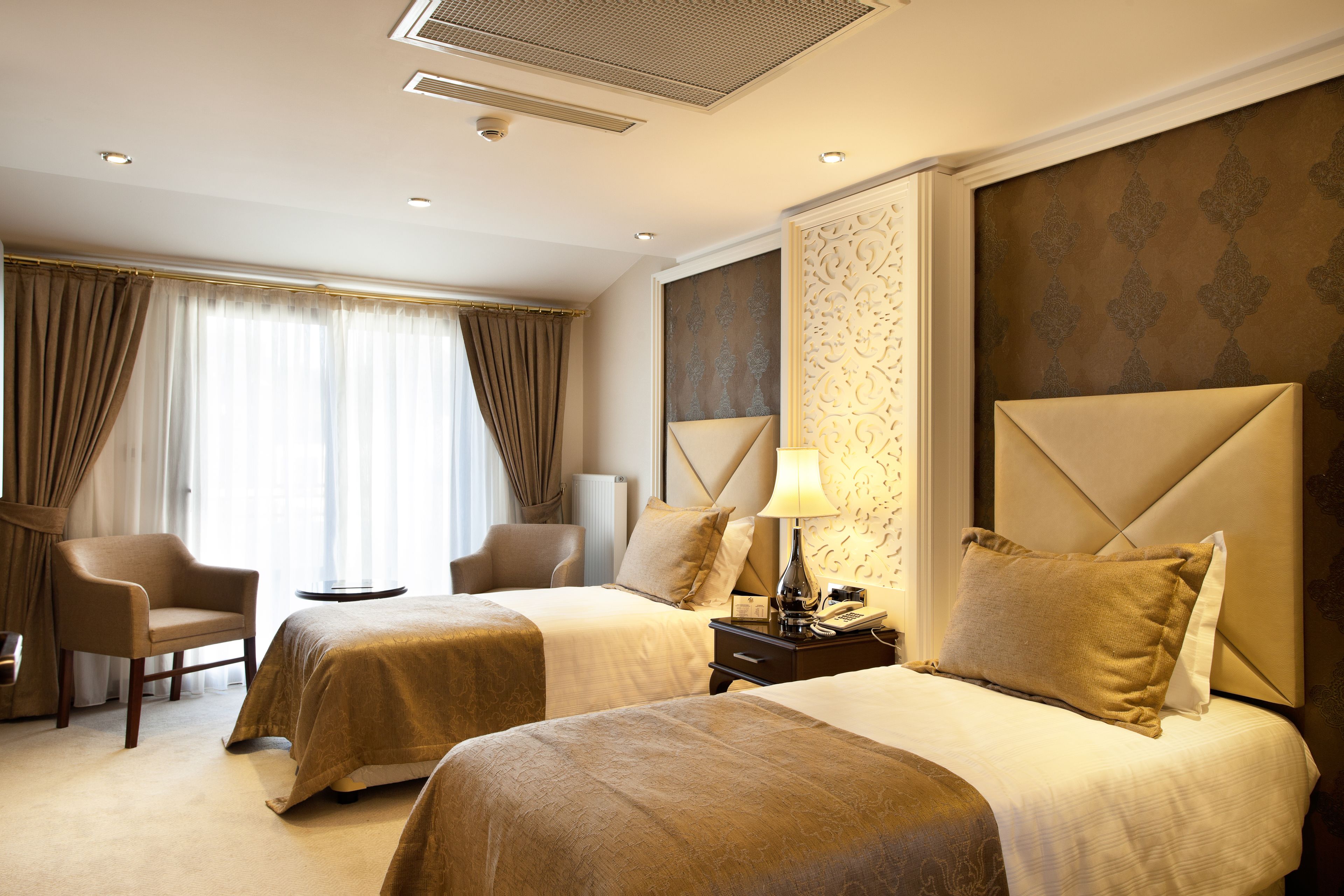 A hotel room with two beds, a sitting area with a chair, a nightstand with a lamp, and a decorative headboard.
