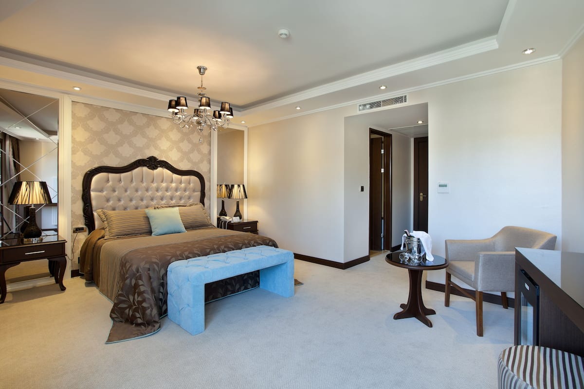 A spacious hotel bedroom with a large bed, a sitting area with a chair and table, and a separate bathroom visible through an open door.
