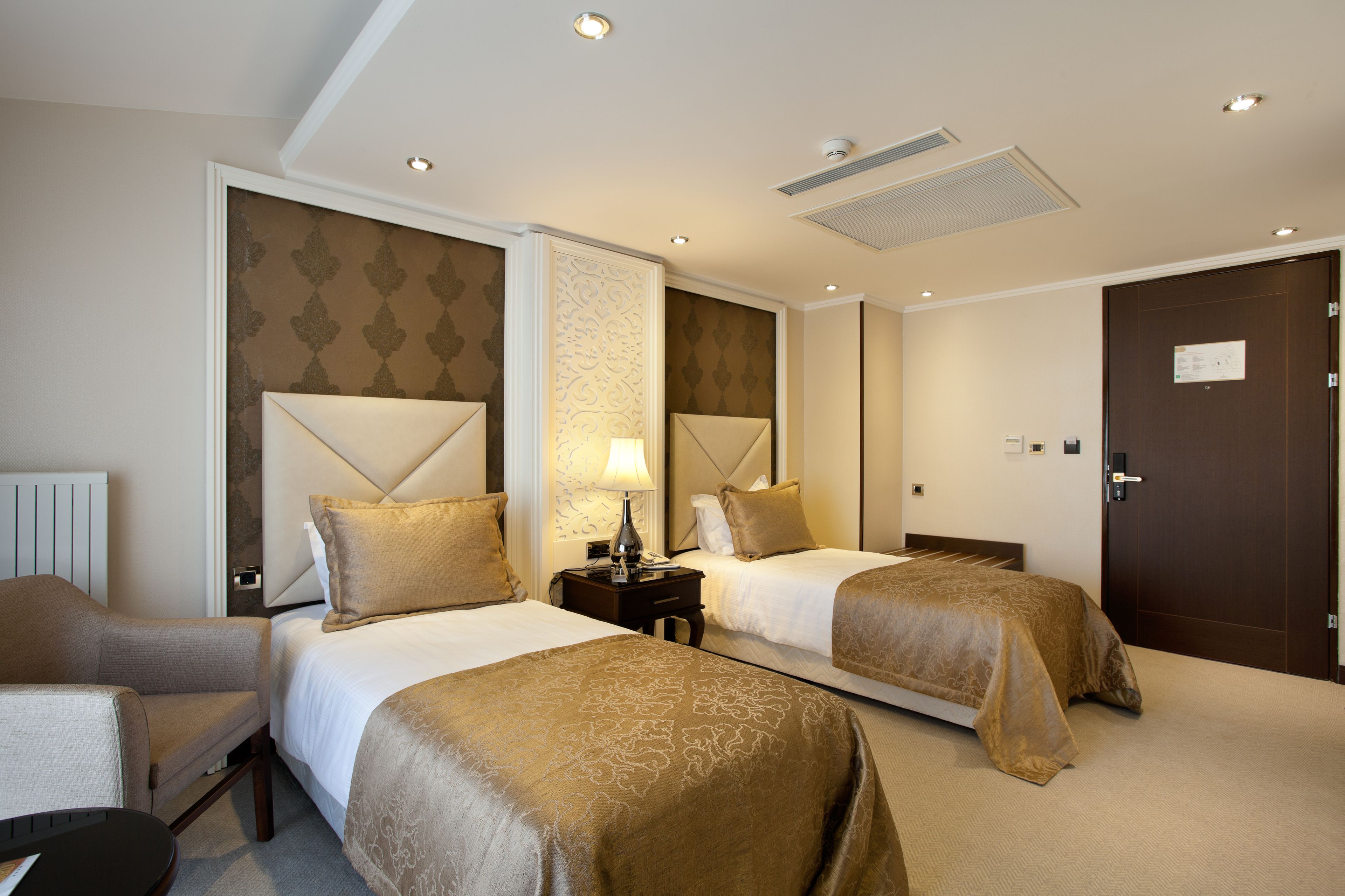 A hotel room with two beds, a nightstand, a lamp, and a chair.