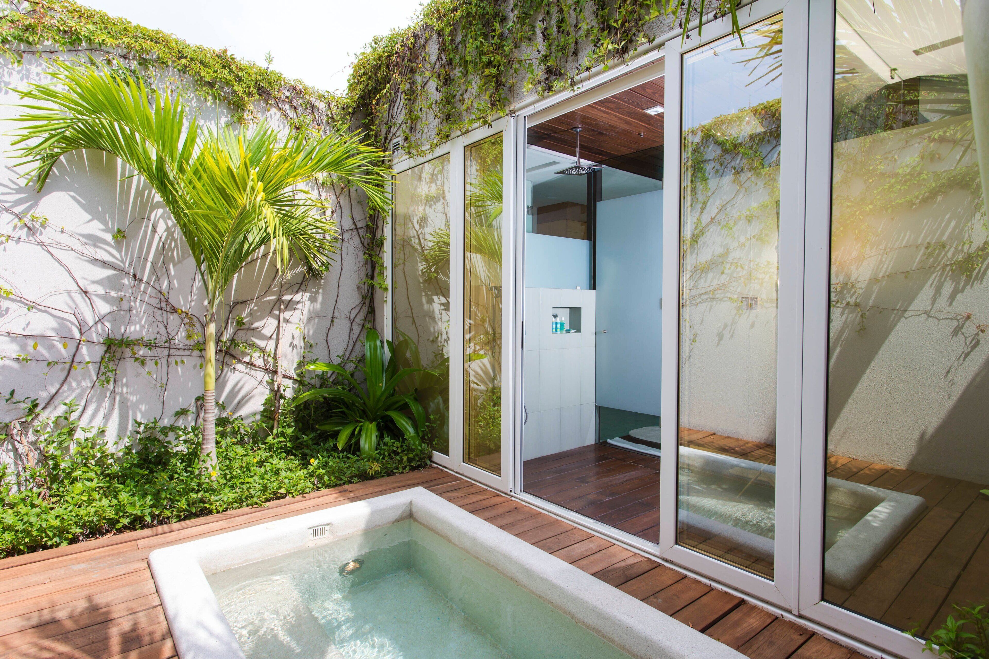 A modern outdoor pool area with a wooden deck, surrounded by lush greenery and a glass-enclosed space.