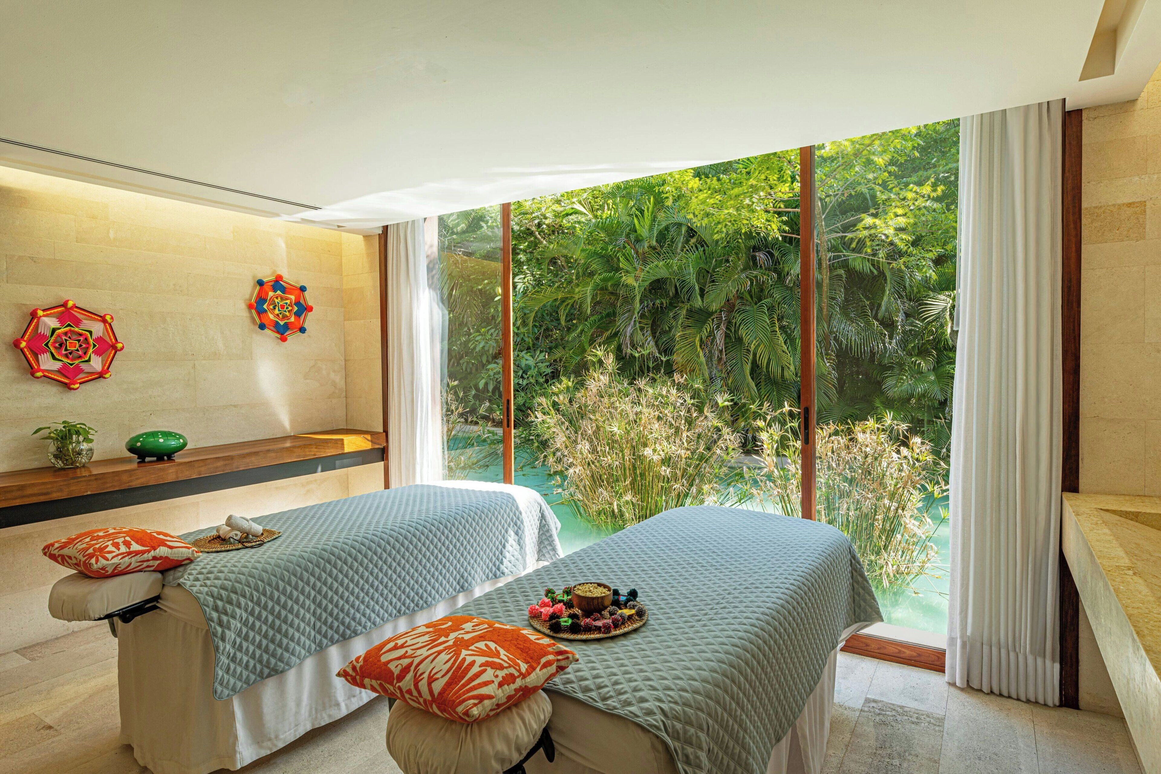 A spa room with two massage tables, a large window with a view of greenery, and decorative wall art.
