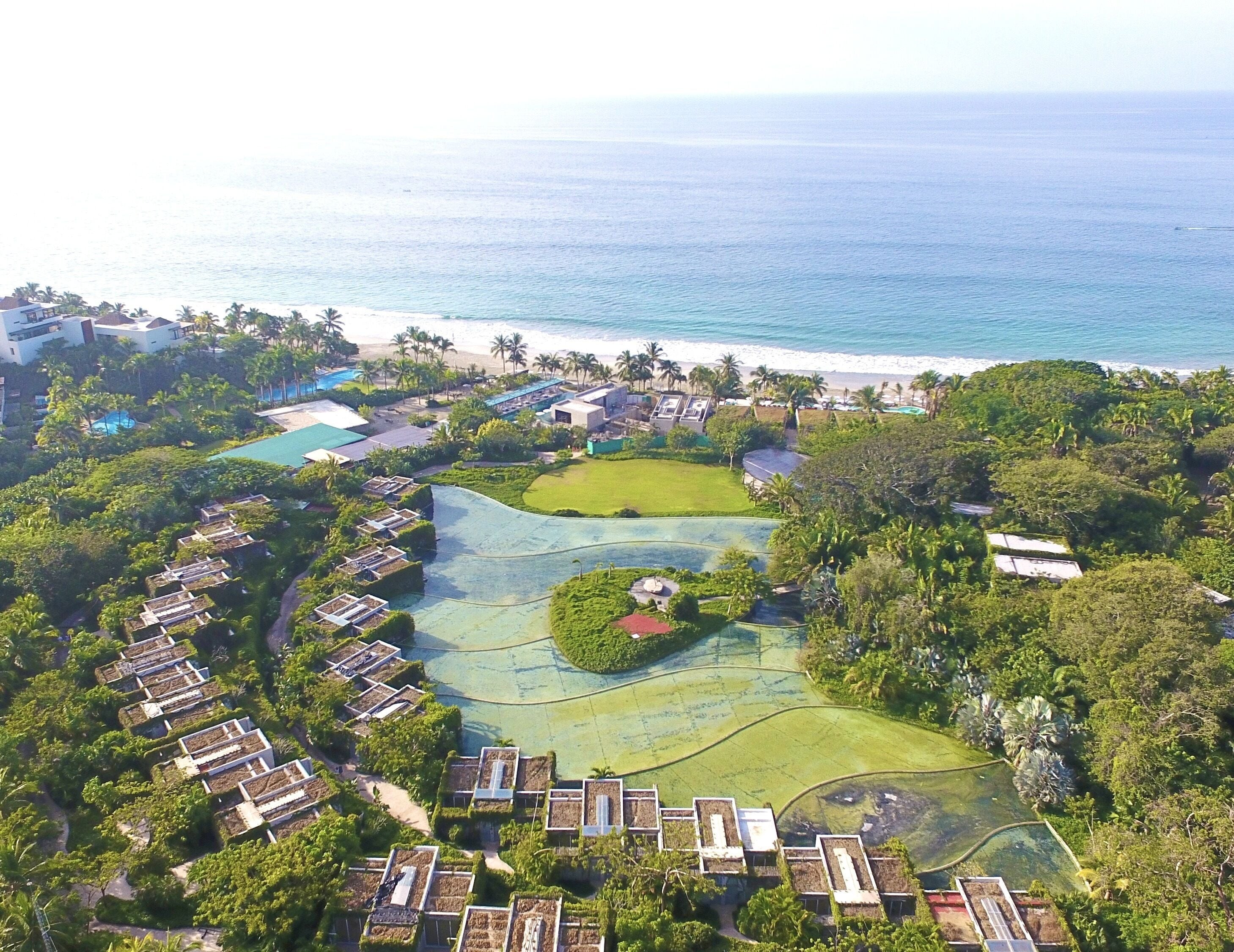 Oversigtsbillede af et resort med flere bungalows, et centralt poolområde og en strand i baggrunden.
