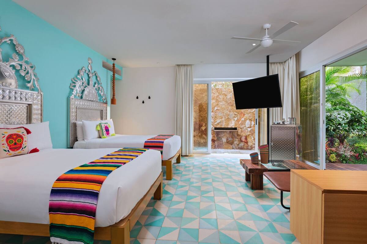 Habitación de hotel con dos camas, televisión, escritorio y balcón con vistas a zonas verdes.