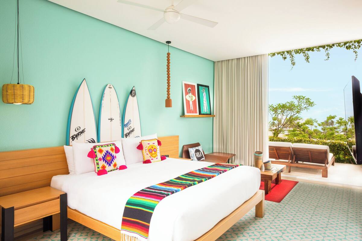 Una habitación de hotel con cama, ventilador de techo, ventana con vistas y balcón con mobiliario.