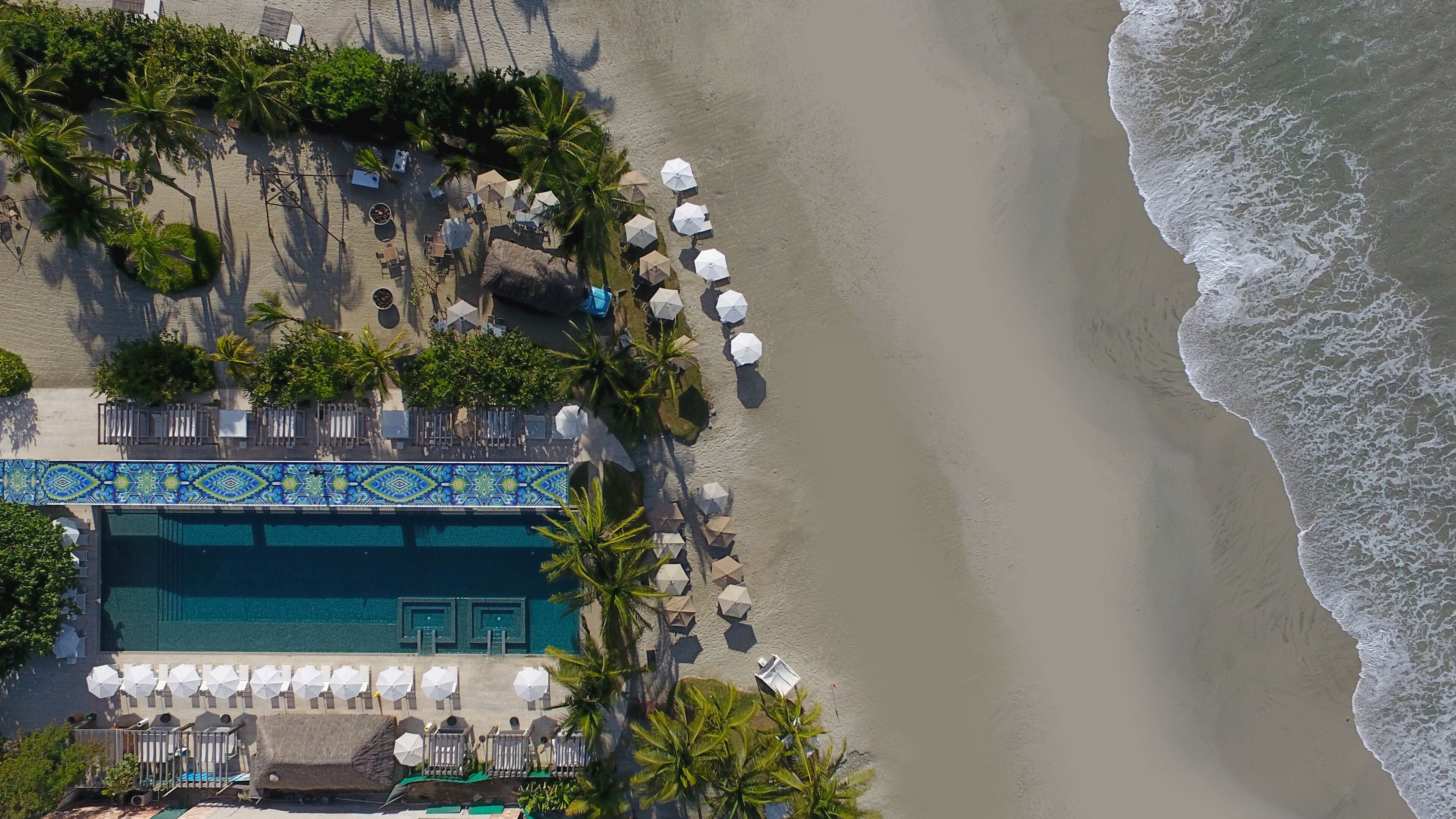 Eine Vogelperspektive auf ein Strandresort mit Pool, Liegestühlen, Sonnenschirmen und Palmen.