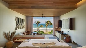 Una habitación de hotel con una cama grande, zona de estar con vista a la playa, televisor de pantalla plana y ventilador de techo.