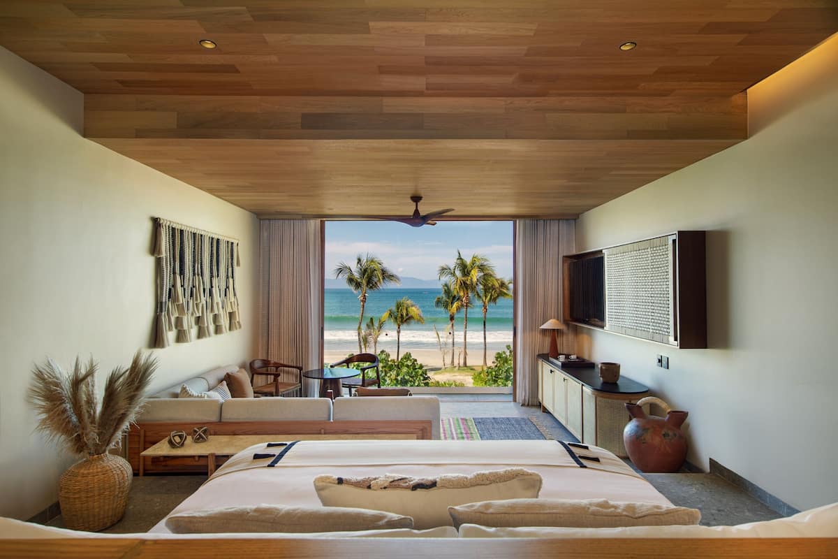 Una habitación de hotel con una cama grande, zona de estar con vista a la playa, televisor de pantalla plana y ventilador de techo.