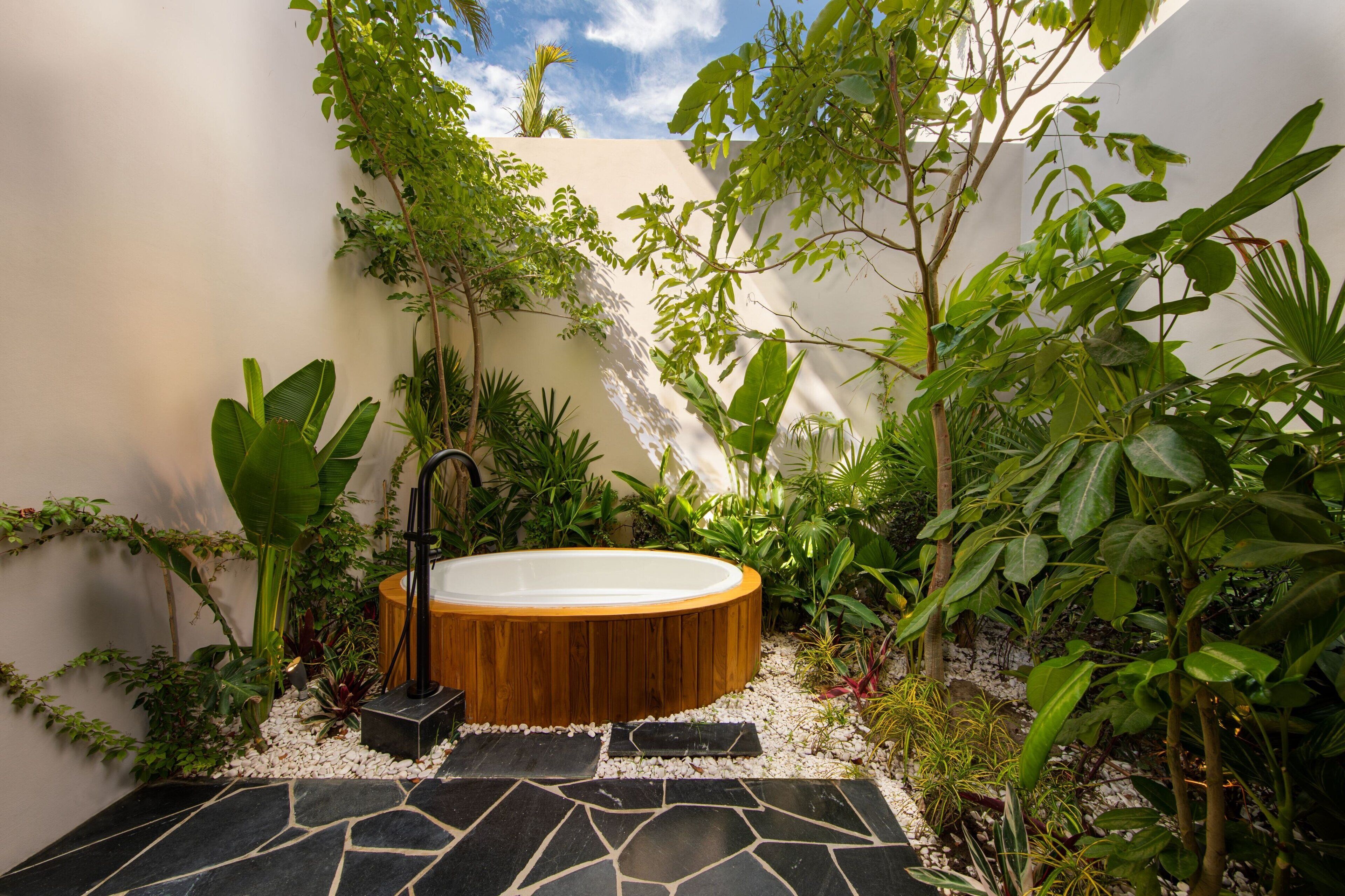 Un espace de bien-être doté d’un bain à remous en bois, entouré d’une végétation luxuriante et doté d’un sol en pierre.
