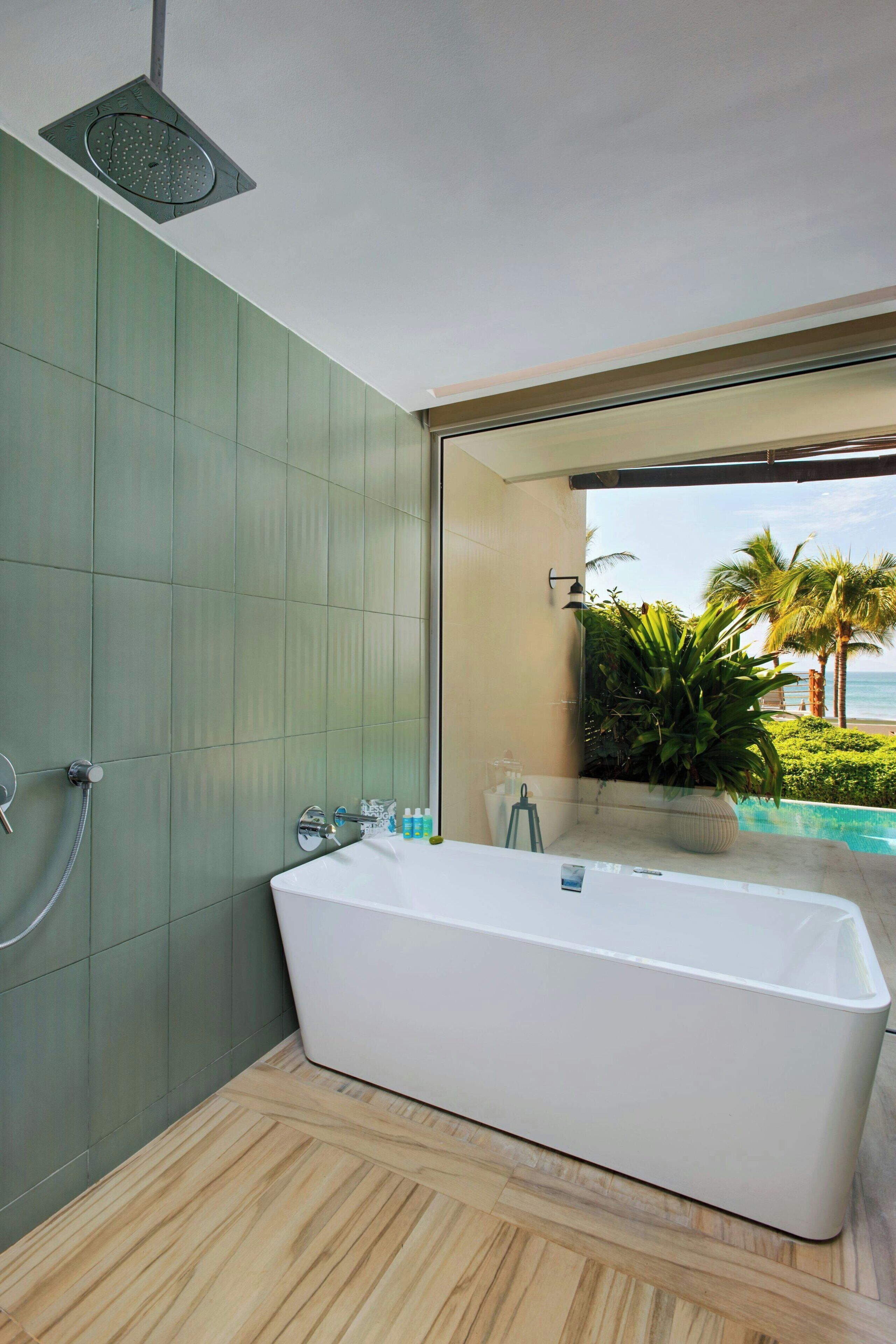 Un baño moderno con una bañera grande, un cabezal de ducha y vistas a una piscina y palmeras.