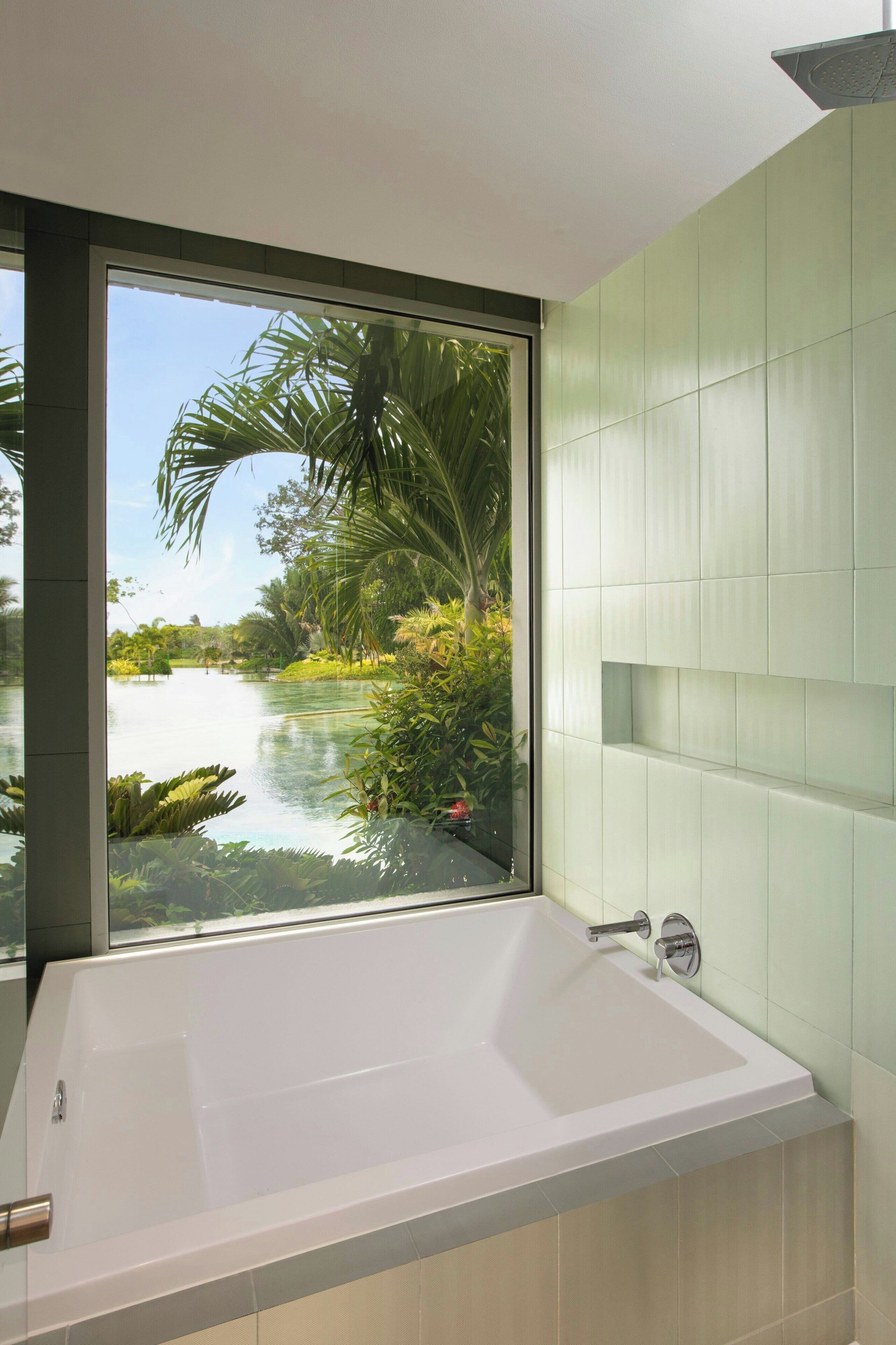 A modern bathroom with a large bathtub, a window offering a view of a lake and greenery, and light green tiled walls.