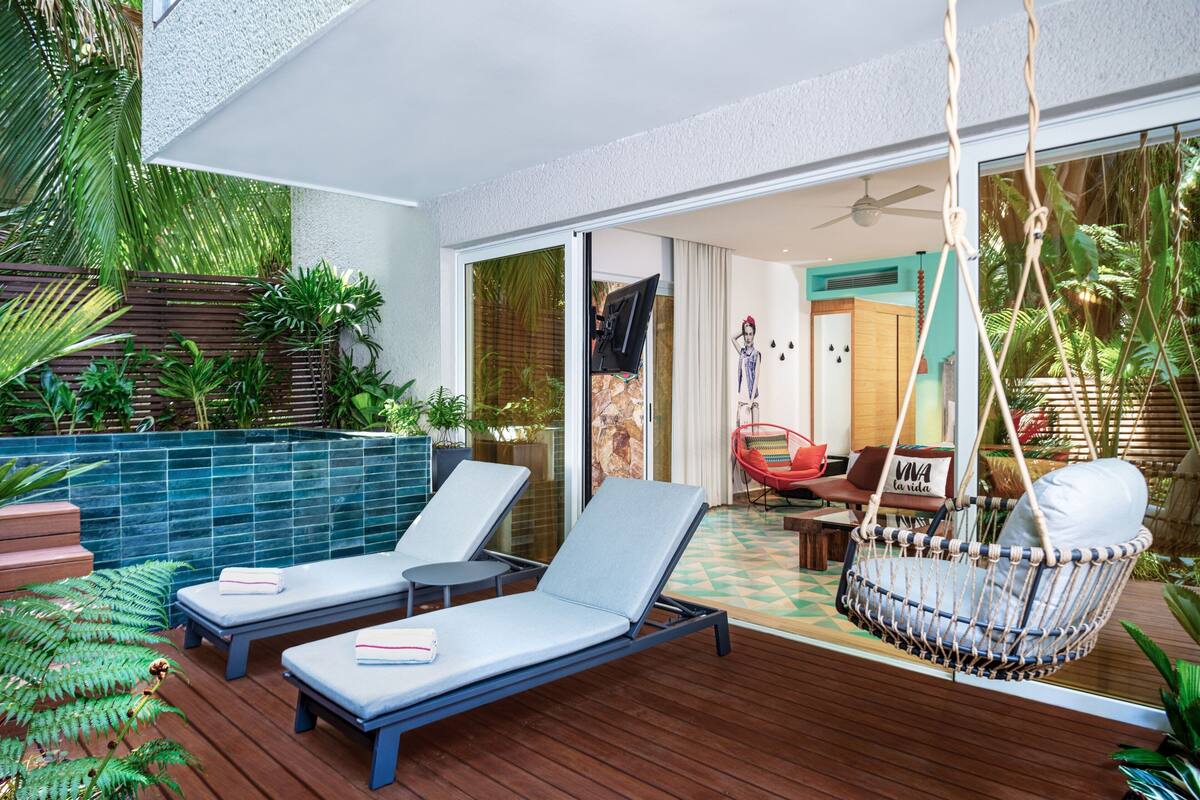 Un área de estar exterior moderna con dos tumbonas, una silla colgante y un ventanal que da a un espacio interior.